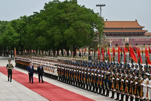 Wide shot showing rows of uniformed soldiers standing to attention as Chinese President Xi Jinping and Russian leader Vladimir Putin walks along a red carpet with Tiananmen Square in the right-hand background.