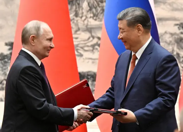 Russian President Vladimir Putin (left) shakes hands with Chinese leader Xi Jinping while both holding red folders of documents with the Chinese and Russian flags behind them.