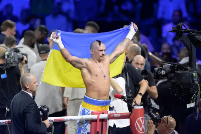 Oleksandr Usyk stands on the side of the ring with a Ukraine flag over his shoulders