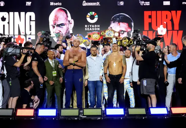 Tyson Fury and Oleksandr stand on a stage with their heavyweight belts in the background