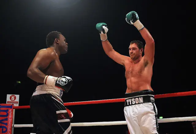 Tyson Fury holds his arms up and stands in front of Derek Chisora