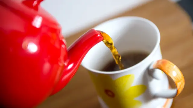 A red teapot pours a drink into a white mug featuring a yellow flower design