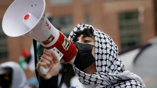 A masked man wearing a pro-Palestinian keffiyeh speaking into a loudspeaer