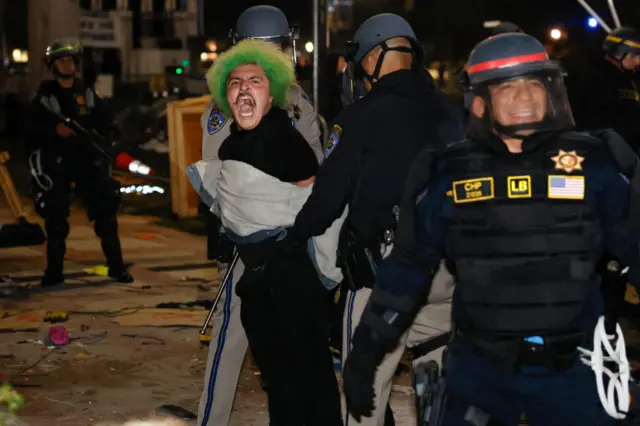 Two police officers wearing helmets arrest a demonstrator with green hair.