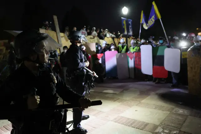 Law enforcement officers wearing riot gear take position and protesters holding shields are standing opposite them.