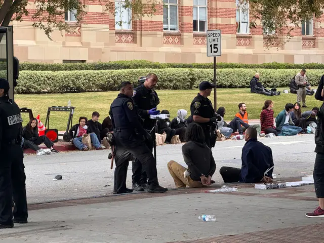 Students with their hands tied behind their backs sit on the pavement