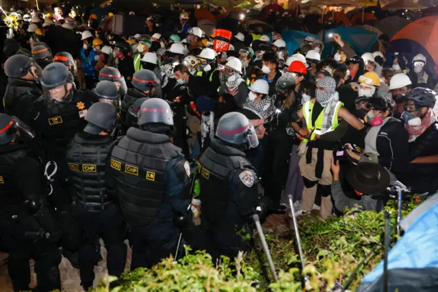 Several police officers in riot gear, standing on the left, face-off with a group of protesters.