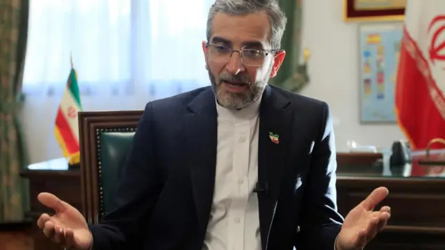 Ali Bagheri Kani sits in a chair and gestures with his hands while an Iranian flag can be seen draped in the background.