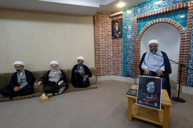 A cleric seated in front of a microphone with a picture of Raisi. Three other clerics are seated to his left in a room. All are offering condolences.