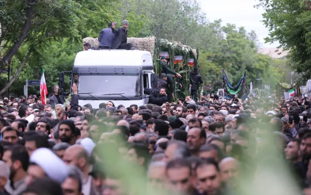 A large crowd moves along with a truck during the funeral procession. Two people can be seen sitting on top of the truck.