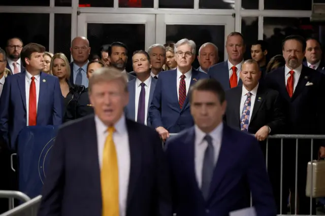 Donald Trump's entourage stands behind him while he speaks to the media