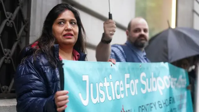 A man and a woman stand holding a sigh which says: "Justice for Subpostmasters"