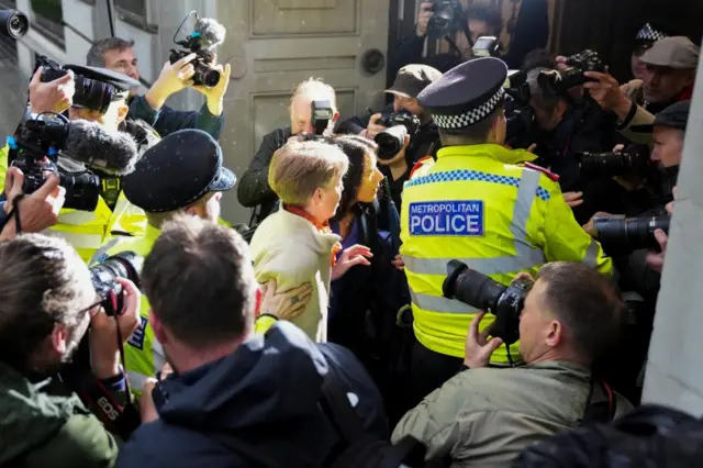 Several photographers stand in front of Vennells with theri cameras raised, as a police officer moves them aside