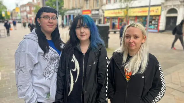 (l to r) Tal, Paige and Dee, three friends, stand together with a generic High Street stretching off behind them in the background. Tal, on the left, has a straight fringe, 50s secretary style glasses with half-rims and long black hair down to their shoulders. They wear a grey zip-up hoody. Paige's hair is a similar length, black on one side and blue on the other half. They're also wearing a black zip-up top. Also in a black zip top is Dee, who has straight shoulder length blonde hair.