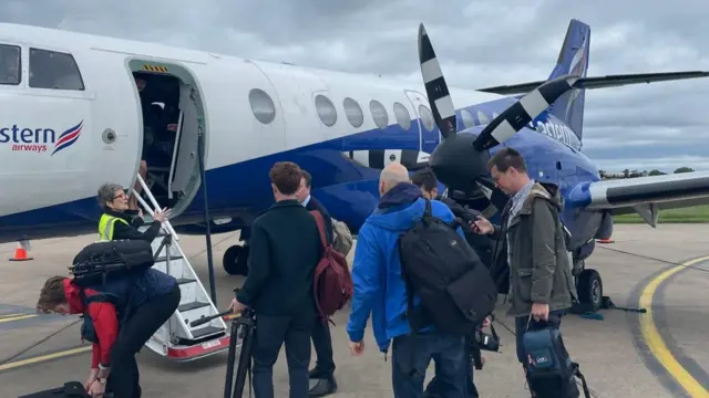 A small plane is seen on a tarmac with a group of journalists and media gathering up their cameras before boarding the small jet liner.