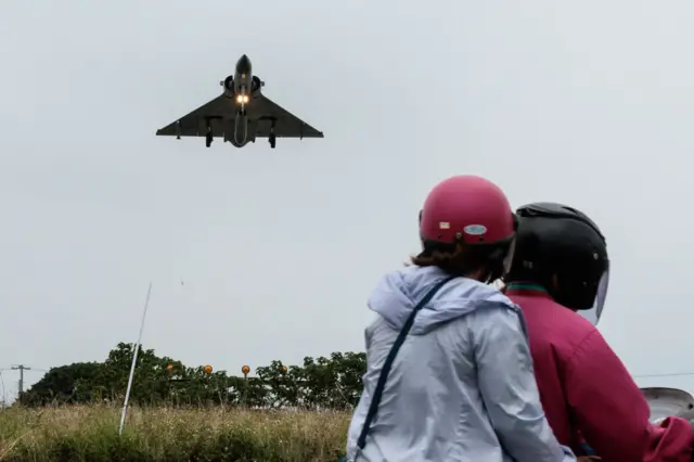 Two people ride a motorcycle as a Taiwanese fighter jet approaches an air force base in Hsinchu