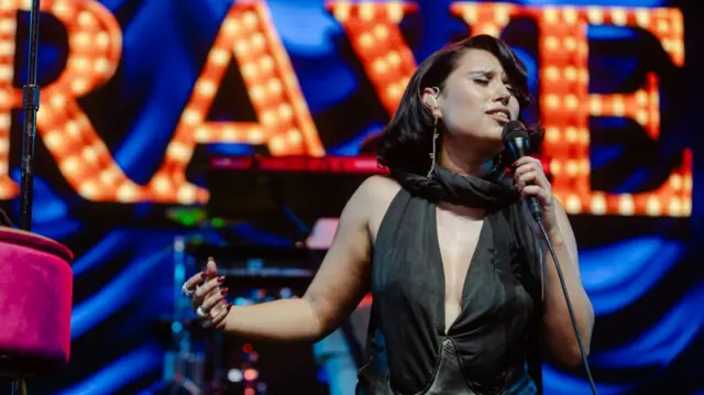 Singer Raye performing on stage, her eyes closed and holding microphone with one arm outstretched. Large sign spelling RAYE behind her.