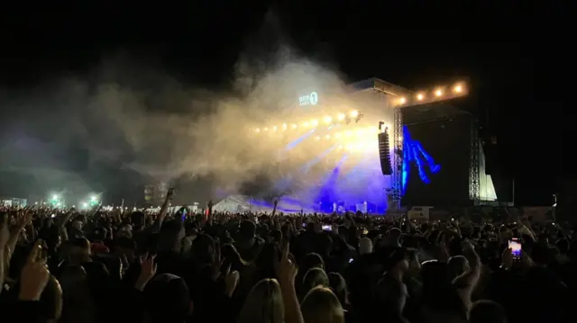 Crowd has their hands in the air in front of yellow and blue light from the Big Weekend main stage