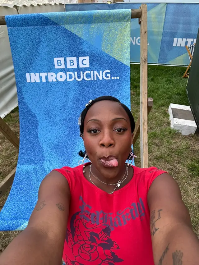 New wave Britpop singer-songwriter Rachel Chinouriri in a bright red top sitting on an oversized deck chair emblazoned with the BBC Introducing logo, taking a selfie and poking her tongue out