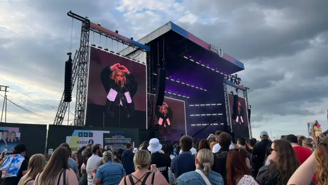 Crowd in foreground with three large screens around a stage, showing Ella Henderson holding hands up in a heart shape