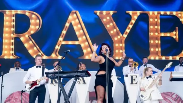 Raye plays Glastonbury. Her name is emblazoned in Gold behind her with an orchestra dressed in white in front of the sign. Raye, a woman, is wearing a black dress singing into a black microphone with her hands raised."