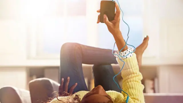 Girl lying down on couch and holding up a music player with headphones in her ears