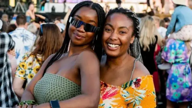 Two young women looking at camera and smiling with large crowd and a stage behind them