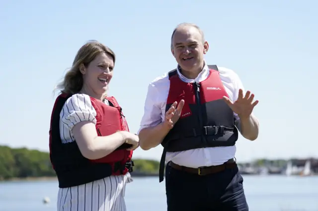 Liberal Democrat parliamentary candidate Jess Brown-Fuller stands next to Liberal Democrat leader Sir Ed Davey near waters in Chichester. Both are wearing red and black life jackets.