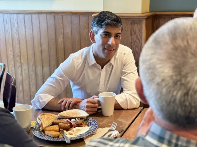 Prime Minister Rishi Sunak holds a cup of tea while sitting at a table with a plate of breakfast in front of him.