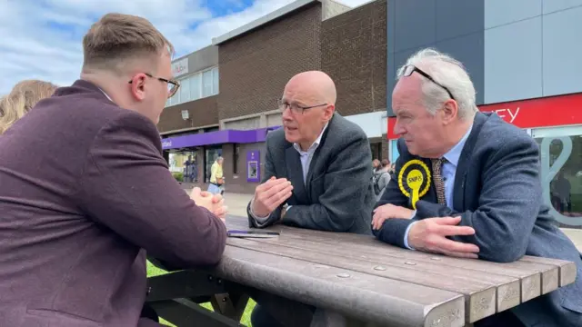 John Swinney sits at a picnic table outside of a shopping centre.