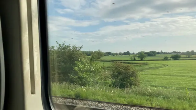 The scene of a field is shown from the perspective of someone travelling inside a train.