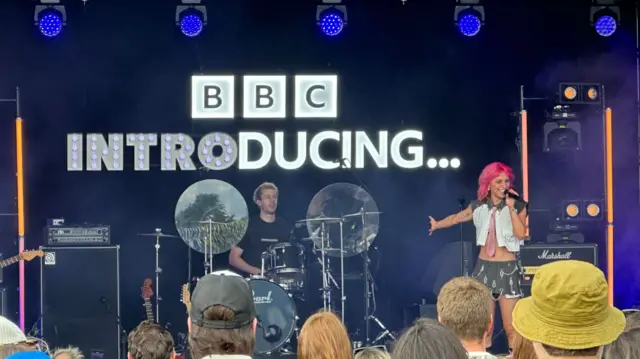 Beth McCarthy, standing on the right hand side of the stage, sings into a microphone, while a man with a drum set is stationed in the middle. There is a screen behind the pair that reads "BBC Introducing".