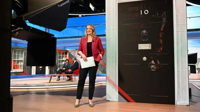 Laura Kuenssberg talks to camera from the studio, with Rachel Reeves and James Cleverly sitting in the background