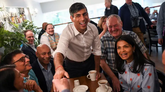 Rishi Sunak extends his hand out to an attendee at the event where he is sitting at table with wife Akshata Murty, Conservative Member of Parliament Bob Blackman and others.