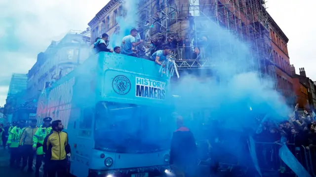 Blue falres surround Manchester City's Ruben Dias holds the Premier League trophy on the bus during the victory parade