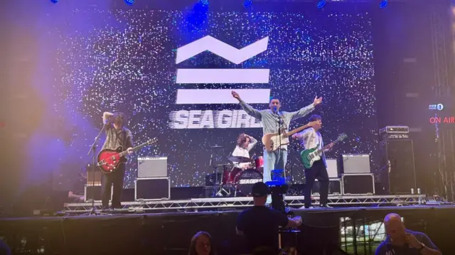 The band Sea Girls on a stage illuminating with blue lights, with a singer holding a guitar standing in the middle and in front of a microphone as two other members pay electric guitars on his side. There is a screen displaying the band's name in the background.