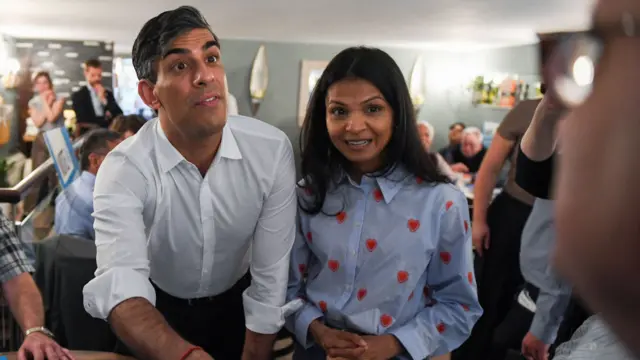 Rishi Sunak stands next to Akshata Murty. He is speaking to another attendee at the event.