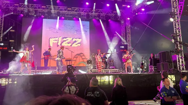 Indie group Fizz performs on a stage with pink and purple lights and a screen reading 'Fizz' in the background.