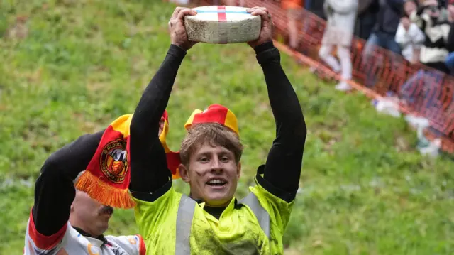 A man wearing a high vis shirt holding a wheel of cheese above his head