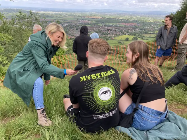 Reporter Georgia Stone pointing a microphone at a man and woman sitting on a hill