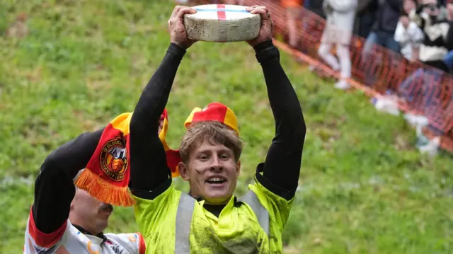 A man wearing a high vis jacket holding a wheel of cheese above his head