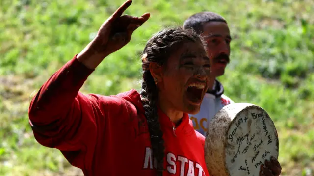 A woman with mud all over her face is holding a wheel of cheese