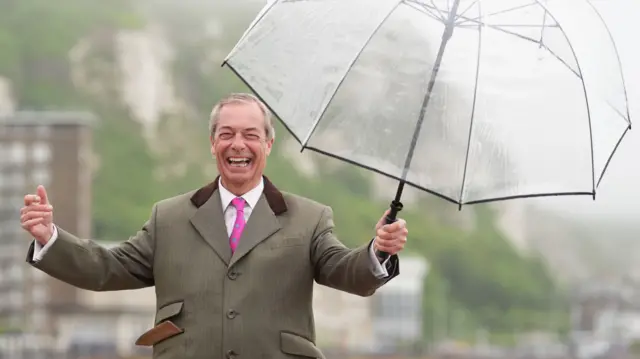 Nigel Farage grins as he holds an umbrella in Dover