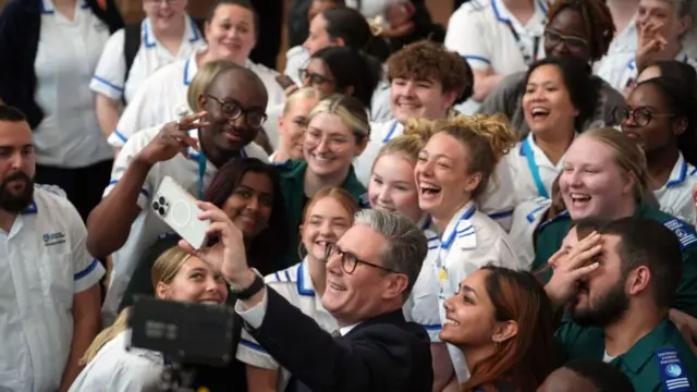 Starmer holds an iphone aloft, smiling for a selfie and medical students gather around him