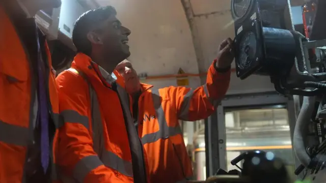 Sunak, in an orange high-vis jacket, smiles as he is shown a gauge on a train