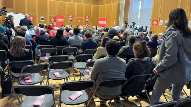 People sit in folding chairs that have been laid out for the audience in a hall. Red Corbyn signs can be seen around the room