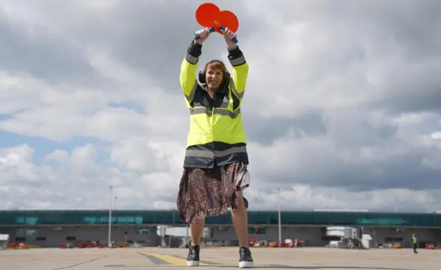 Rayner, dressed in a yellow high-vis jacket, stands on tarmac holding two red paddles in a cross over her head