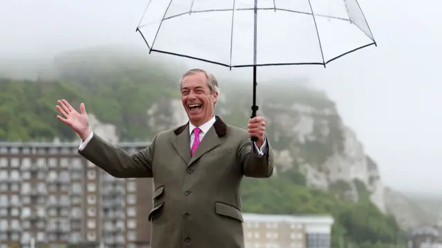 Nigel Farage holding a see through umbrella and grinning with the White Cliffs of Dover in the background