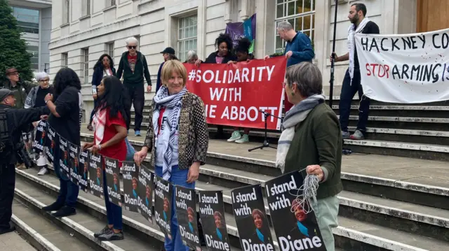 Roughly a dozen people stand on the town hall steps, some wearing Palestinian keffiyehs, with signs in support of Abbott