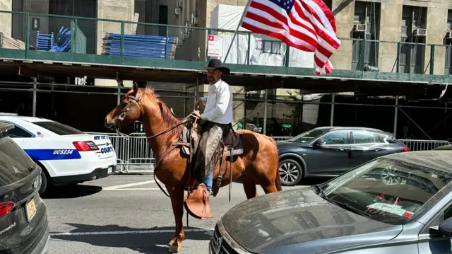 A man on horse holds an american flag in the middle of a busy street